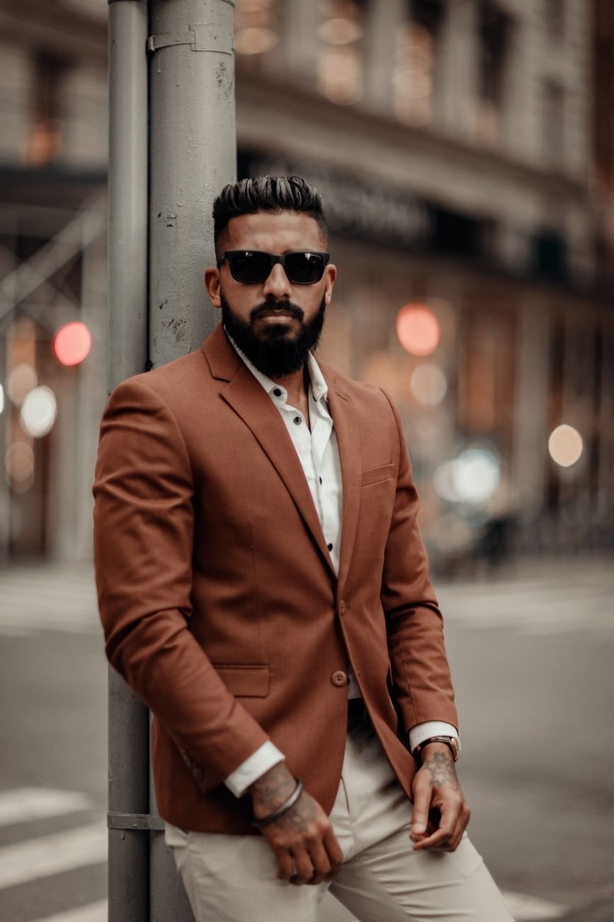 Man with sunglasses posing elegantly in an urban setting wearing a brown blazer